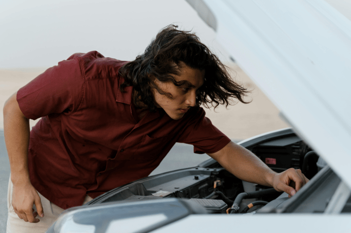 A young man in a maroon shirt leaning over an open car hood, inspecting the engine.