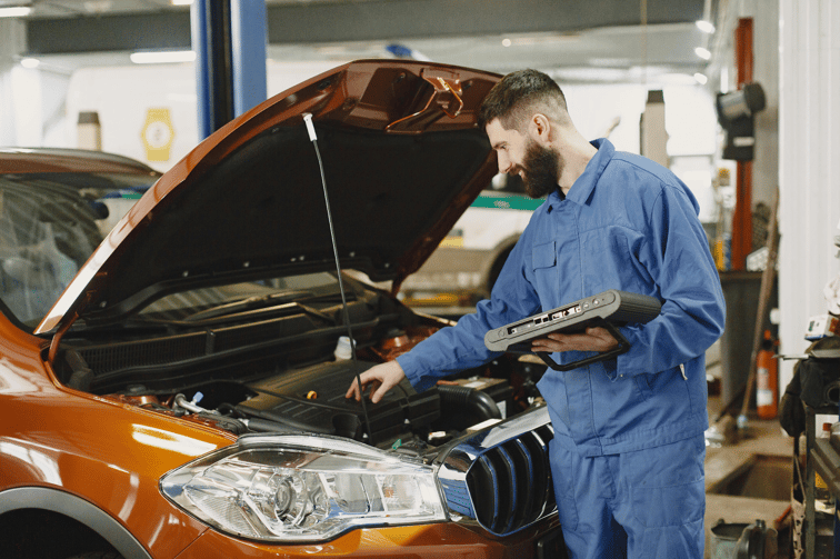 A smiling mechanic in blue coveralls holding a diagnostic tool while inspecting an orange car's engine.