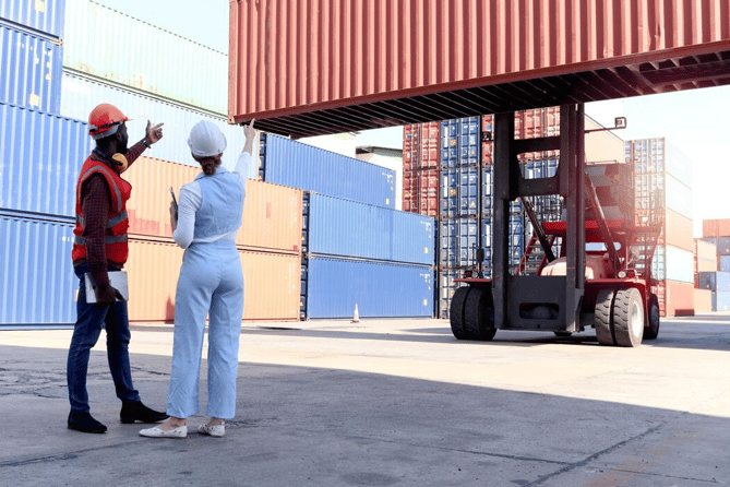 Two port workers in hard hats are pointing at a shipping container being lifted by a forklift.