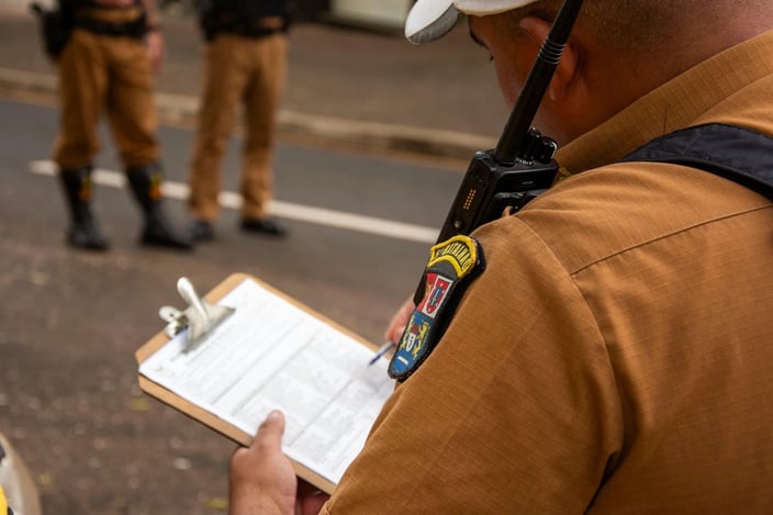 An officer in uniform writes on a clipboard while another stands in the background.