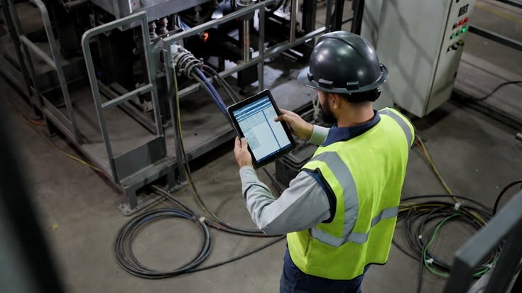 A field worker in a manufacturing facility using a tablet to complete a digital compliance checklist on the production floor.