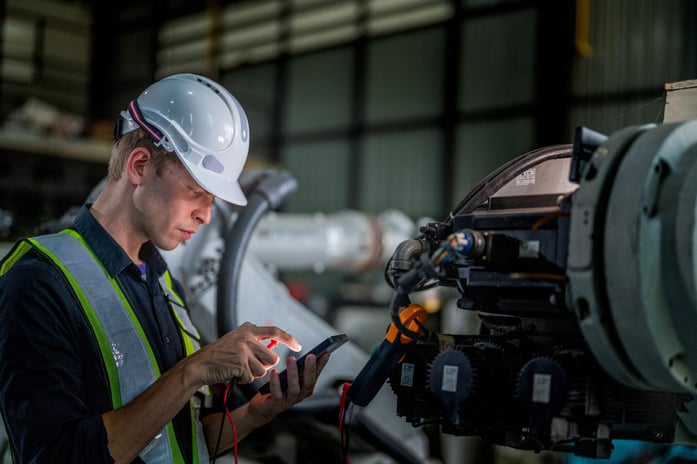 An engineer in a hard hat and safety vest is inspecting industrial machinery with a diagnostic tool.