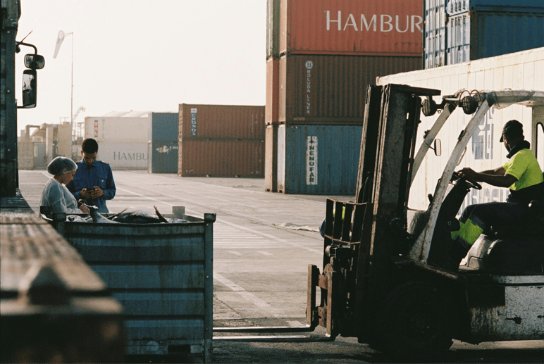 Workers and a forklift operator at a shipping port with stacked colorful cargo containers.