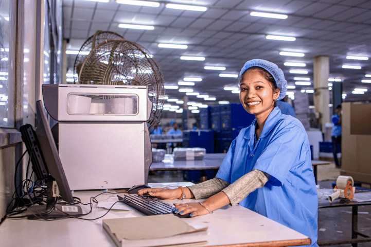 Smiling woman in a blue hairnet and lab coat working at a computer in a factory.