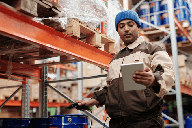 Warehouse worker in a blue beanie holding a tablet and a barcode scanner near storage shelves.