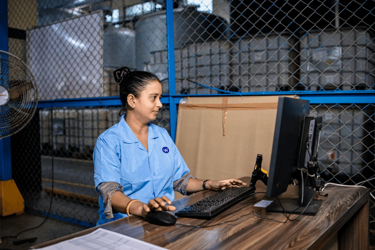 A factory worker in a blue uniform operating a computer in a warehouse.