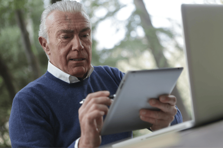 Older man in a blue sweater reading a tablet while sitting near an open laptop outdoors.