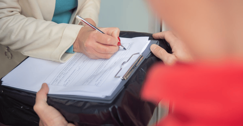 Person filling out a form on a clipboard while another person watches nearby.