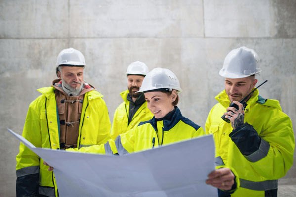 Four workers wearing white hard hats and yellow safety jackets review construction plans at a worksite.