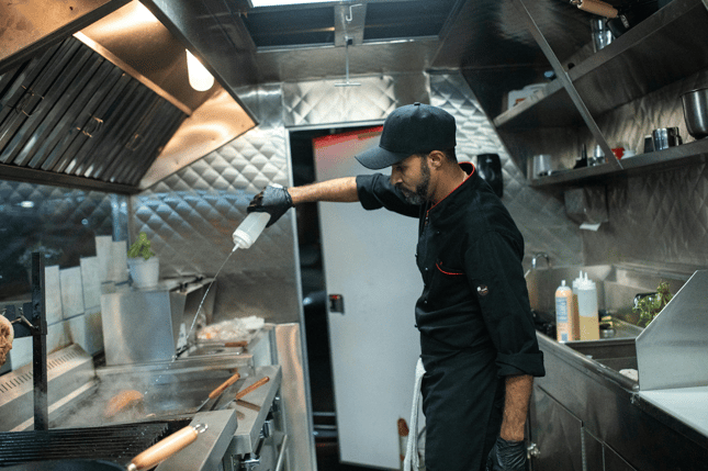 Chef in a black uniform and cap cleaning a commercial kitchen exhaust hood in a professional restaurant kitchen.
