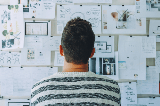 Rear view of a person in a striped sweater studying a wall covered with documents, diagrams, photos, and planning materials organized on a whiteboard.