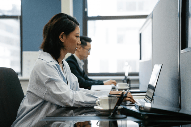 Woman in business attire working at a laptop in a modern office with a colleague in the background.