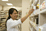 A smiling pharmacist in a white coat reaches for medication on a shelf in a well-lit pharmacy