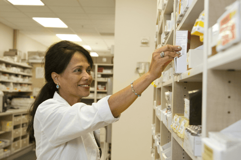 A smiling pharmacist in a white coat reaches for medication on a shelf in a well-lit pharmacy.
