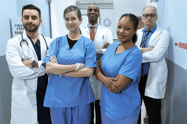 Group of five healthcare professionals, including doctors in white coats and nurses in blue scrubs, standing together in a hospital corridor.