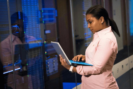 Professional woman in a pink shirt using a laptop in a server room with blue-lit server racks in the background.