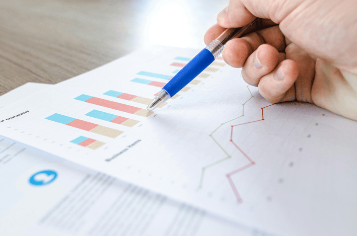 Hand holding a blue pen pointing at printed business charts with colorful bar graphs and line graphs.