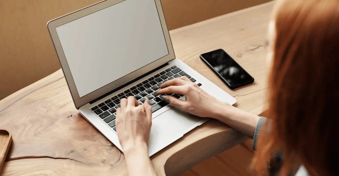 Person typing on laptop at wooden desk with smartphone nearby.