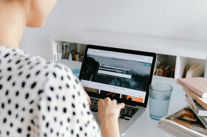 Person in polka dot shirt browsing stock photo website on laptop with water glass and books nearby.