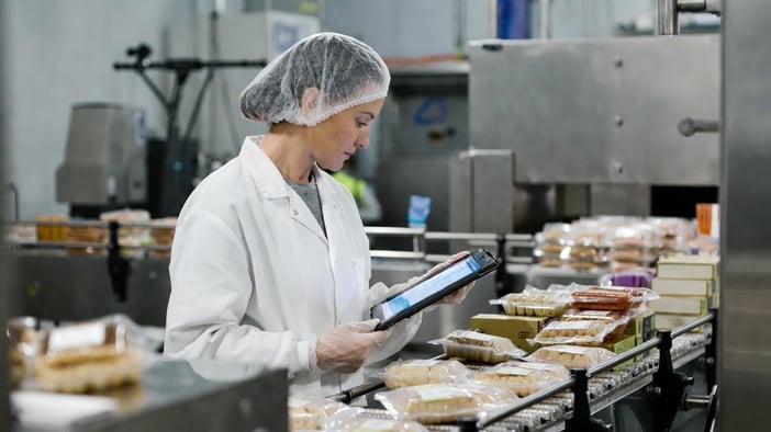 Quality control manager examining packaged food products while holding a tablet in a food processing facility.