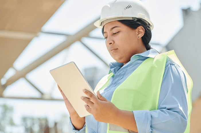 Mulher com capacete e colete de segurança usando um tablet em uma construção ou instalação industrial.