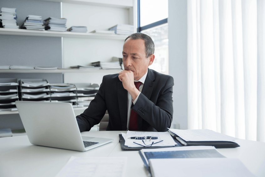 Businessman in dark suit and red tie working thoughtfully on laptop at modern office desk.