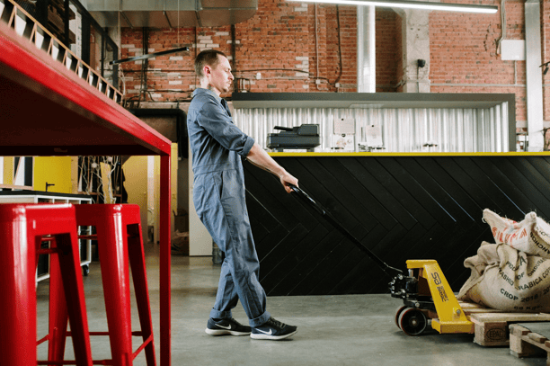 Worker in coveralls operating a yellow pallet jack in an industrial warehouse space with exposed brick walls.