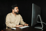 Man in beige shirt working at desktop computer with keyboard and mouse against a dark background.