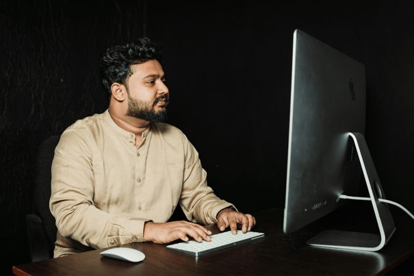 Man in beige shirt working at desktop computer with keyboard and mouse against a dark background.