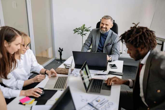 Four business professionals collaborating at a table with laptops in a modern office setting.