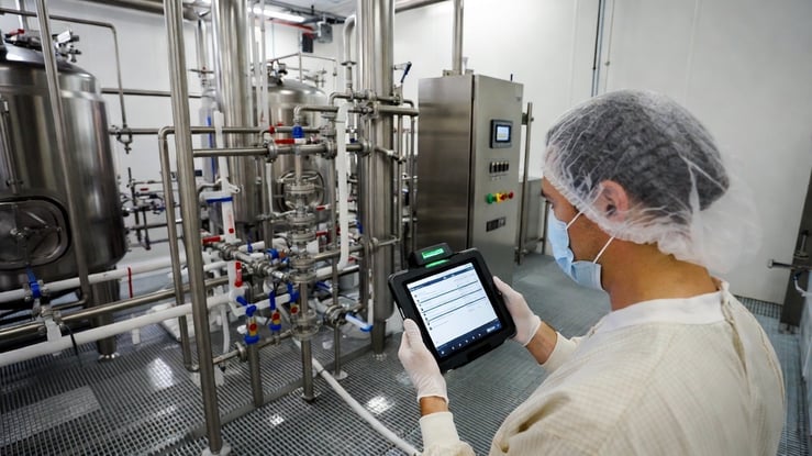 A food and beverage manufacturing plant floor with a worker using a mobile tablet to complete a digital inspection checklist.