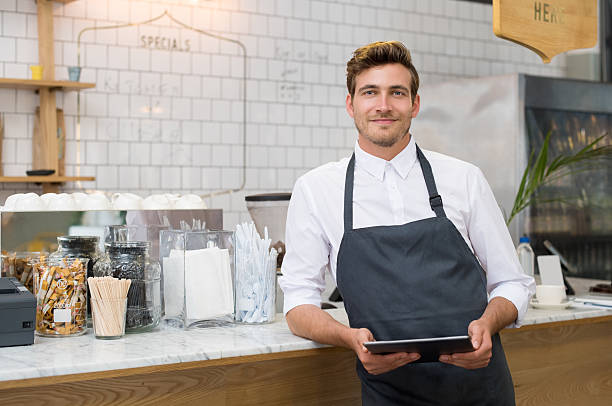A smiling café worker in a dark apron holding a tablet behind a marble counter.