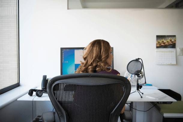 A person with curly brown hair sitting in an ergonomic office chair at a desk, working at a computer monitor, viewed from behind.