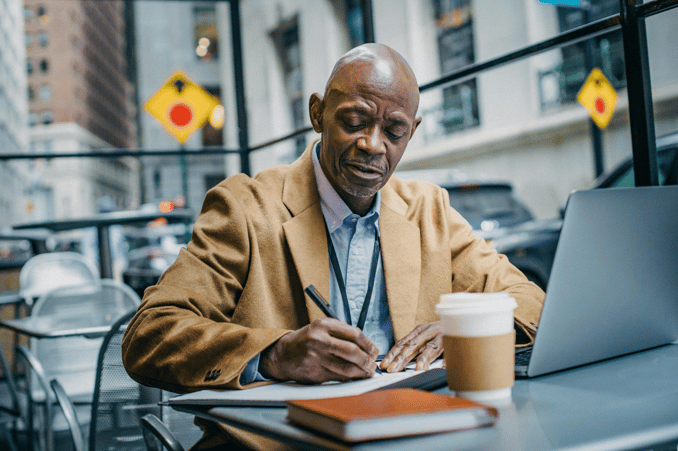 A man in a tan blazer and blue shirt writing notes on paper at an outdoor café table, with a laptop and coffee cup nearby in an urban setting.