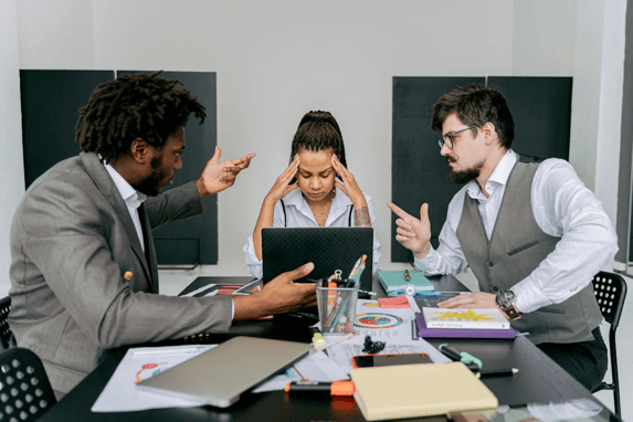 A stressed woman holds her head while two male colleagues argue animatedly during an office meeting.