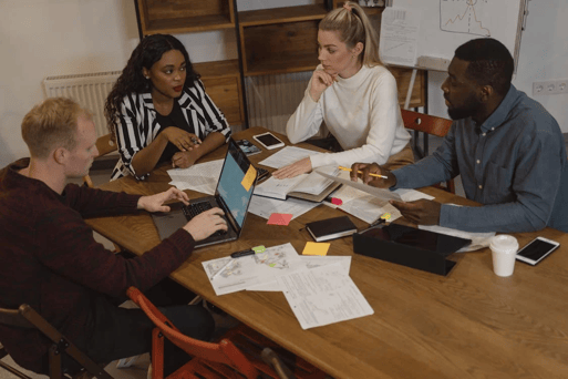 Four colleagues collaborate around a wooden table covered with laptops, documents, sticky notes, and notebooks.