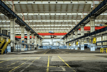 Interior of a large industrial manufacturing facility with concrete columns, overhead cranes, and yellow floor markings.