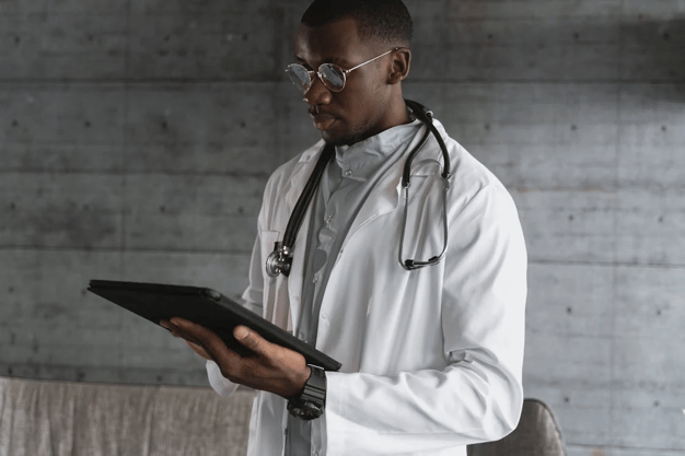 Doctor in white coat with stethoscope and glasses reviewing information on a tablet device.