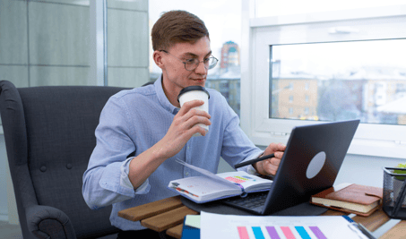 Man with glasses working on a laptop while holding a coffee cup at a desk.