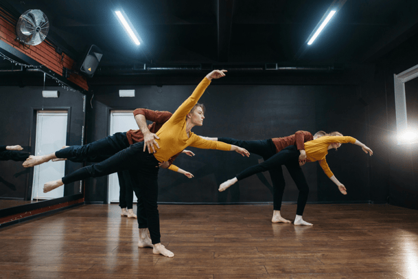 Four contemporary dancers perform synchronized leaning poses in a dark studio with wooden floors and mirrors.