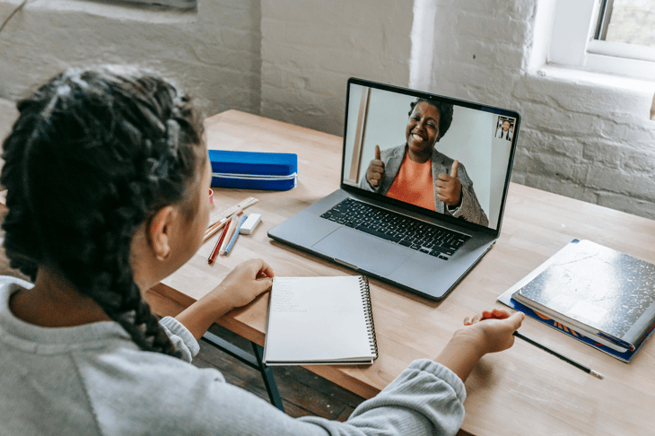 Young student with braided hair attending a video call on a laptop with teacher giving thumbs up on screen.
