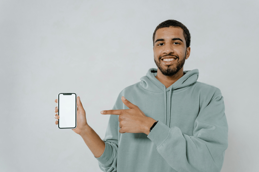 Smiling man in a gray-green hoodie holding a smartphone with a blank screen and pointing at it.