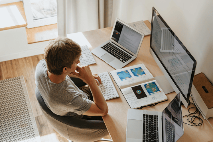 Person working at home office desk with dual monitors, laptops, and notes.