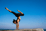Woman performing yoga handstand pose on coastal rocks against clear blue sky and ocean.