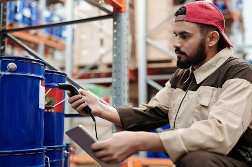 Warehouse worker wearing a red cap scans a barcode on blue industrial drums while holding a tablet.