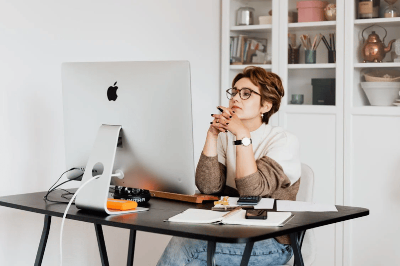 Woman with short hair and glasses sits thoughtfully at a desk with an iMac in a home office.