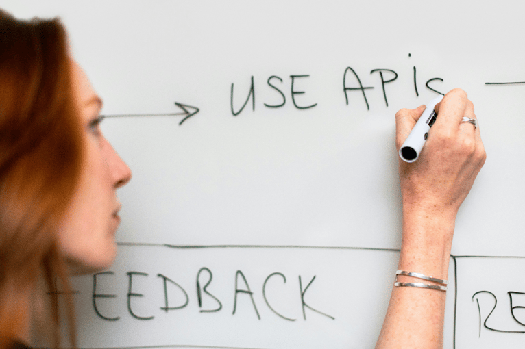 Person writing "USE APIs" on whiteboard with marker, wearing bracelet on wrist.