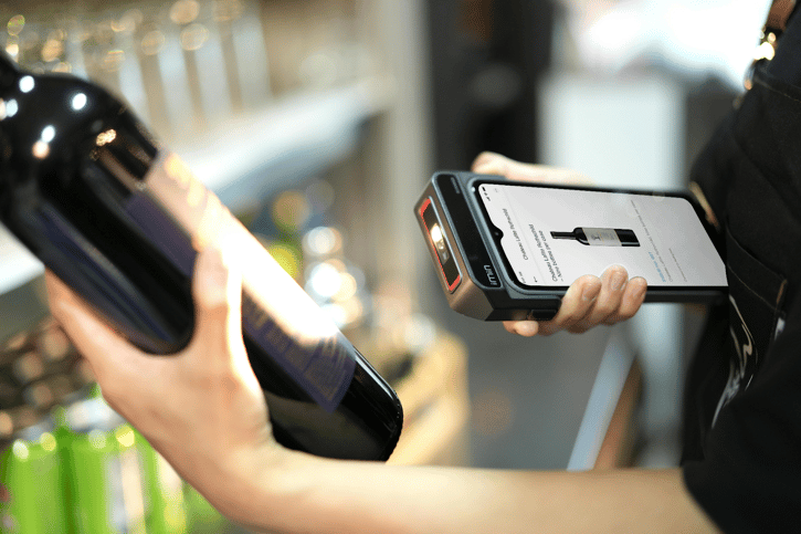 Hand scanning a wine bottle with a handheld device displaying product information on its screen.