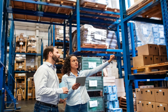 Two warehouse workers with a tablet reviewing inventory on blue industrial shelving stocked with boxes. (Image courtesy of The Candidate Source)