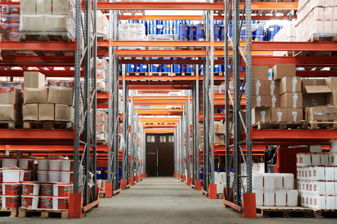 Long warehouse aisle with orange steel shelving stocked with boxes, blue drums, and plastic containers.